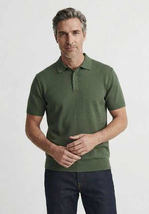 Homme aux cheveux gris portant un polo vert à manches courtes et un jean foncé, debout les mains jointes devant un fond uni.