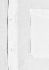 Gros plan sur le tissu d'une chemise blanche montrant les boutons avant et une poche poitrine sur le côté droit.