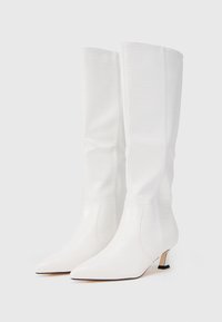 Botas altas de cuero blanco con textura de cocodrilo, con punteras afiladas y tacones kitten bajos sobre un fondo liso.