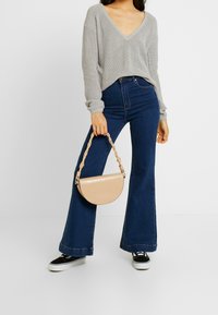 Pull en maille gris avec col en V, jean bleu évasé et sac à main beige en demi-lune avec une finition texturée et une bandoulière tressée. Baskets noires.