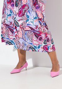 Zapatos de tacón alto de cuero rosa con un diseño de punta afilada descansando sobre una superficie clara, combinados con un vestido de estampado paisley colorido.