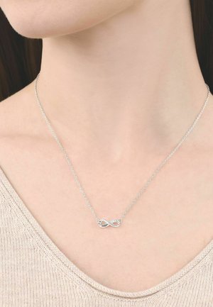 Collar delicado con símbolo de infinito en plata sobre una cadena fina, llevado alrededor del cuello de una persona que lleva un suéter beige de punto.
