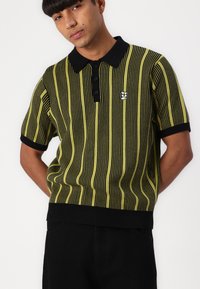 Knitted polo shirt in green and black vertical stripes, featuring a black collar, three button placket, and a small logo on the chest.