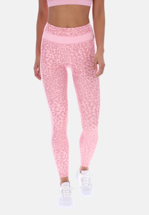 Γυναίκα που φορεί ροζ κολάν με λεοπάρ μοτίβο ψηλής μέσης και ασορτί αθλητικό μπουστάκι, σε συνδυασμό με λευκά αθλητικά παπούτσια, στέκεται μπροστά σε μονοχρωμο φόντο.