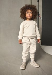 Kreemivärvi dressipluus ja dressipüksid, mis on valmistatud pehmest kangast, soonikkoes varrukate ja nööriga vöökohaga. Kandmisel heledate värvidega vabaajajalatsid.