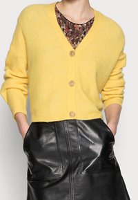 Cárdigan amarillo de punto con escote en V, que cuenta con un cierre de botones y textura acanalada, combinado con una falda de cuero negra.