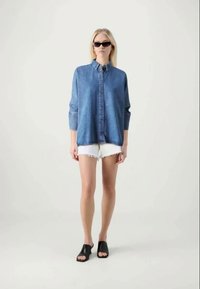 Helles, blaues Denim-Hemd mit langen, hochgerollten Ärmeln, kombiniert mit weißen, ausgefransten Shorts und schwarzen Rutsch-Sandalen.