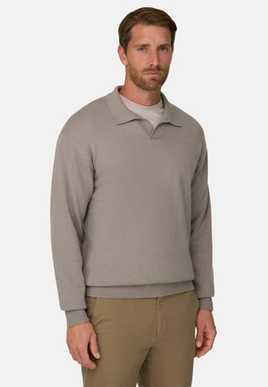 Un homme porte un pull beige en maille à col par-dessus une chemise blanche avec un pantalon olive devant un fond clair uni.