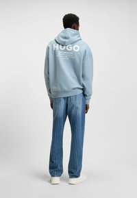 Lichtblauwe hoodie met witte "HUGO" logo, gecombineerd met blauwe gestreepte jeans en witte sneakers, wat een ontspannen, casual uitstraling benadrukt.