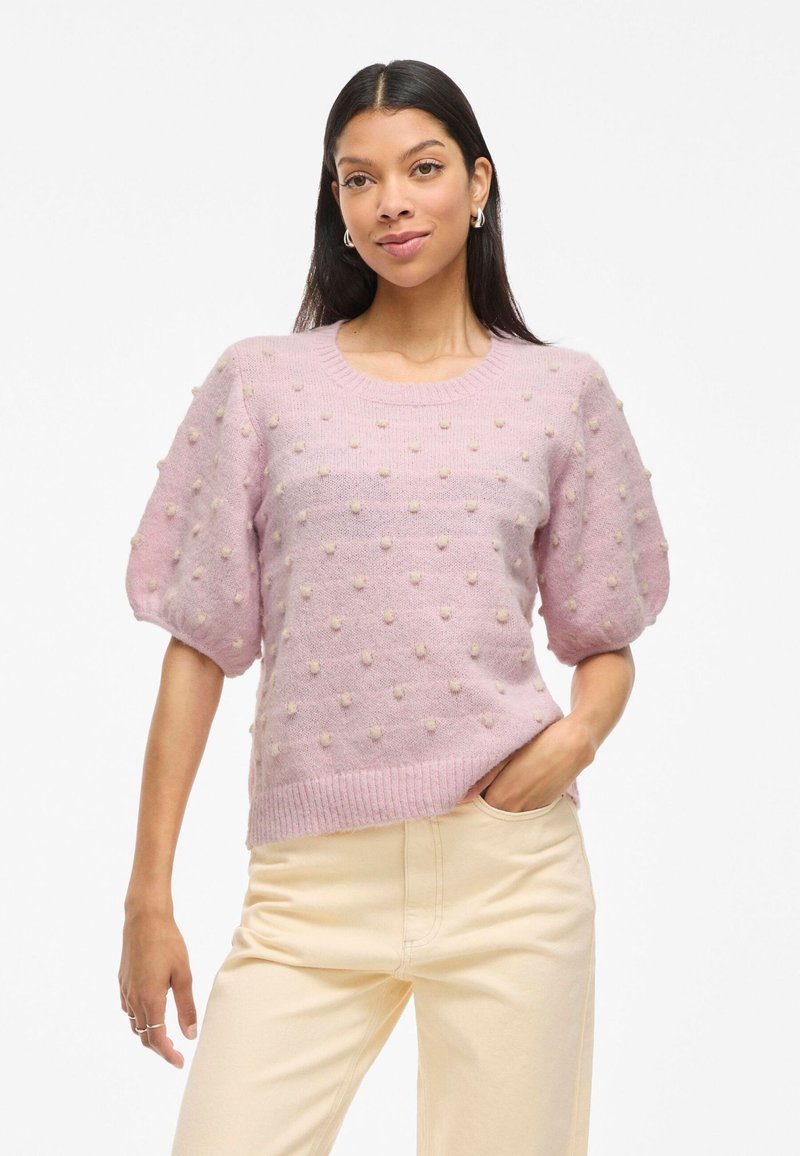 Mujer vistiendo un suéter de punto rosa claro con detalles texturizados de pompón y mangas abullonadas, combinado con pantalones crema de cintura alta.