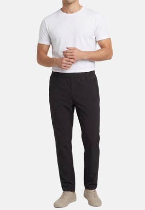 Hombre con una camisa blanca de manga corta lisa, pantalones negros con cintura elástica y zapatos casuales beige, de pie con las manos entrelazadas.