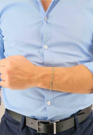 Braccialetto color oro con un design intrecciato complesso, indossato al polso, abbinato a una camicia azzurra con bottoni e una cintura scura.