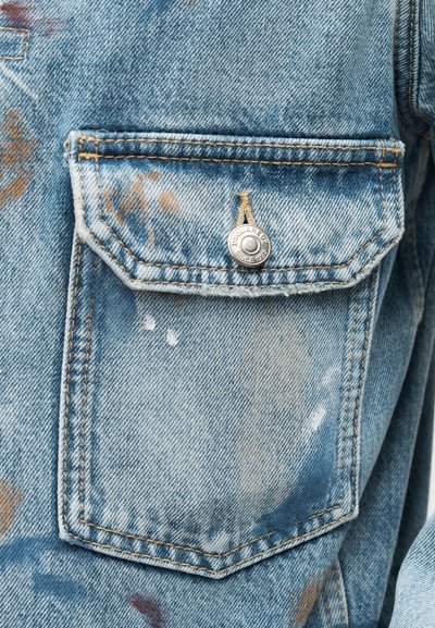 Τσέπη ανοιχτό μπλε τζιν μπουφάν με πορτοκαλί ραφές, μεταλλικό κουμπί με χαραγμένο «Dsquared Since 1964» και διακριτικές καφέ και λευκές πιτσιλιές μπογιάς.