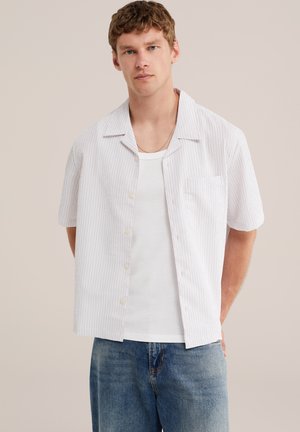 Jeune homme portant une chemise blanche à fines rayures à manches courtes sur un t-shirt blanc et un jean bleu, debout les mains derrière le dos.