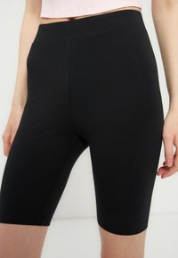 Γυναίκα που φοράει μαύρα στενά σορτς μέχρι το γόνατο με ψηλή μέση και κοντό rib top σε ροζ χρώμα, στέκεται μπροστά σε απλό λευκό φόντο.