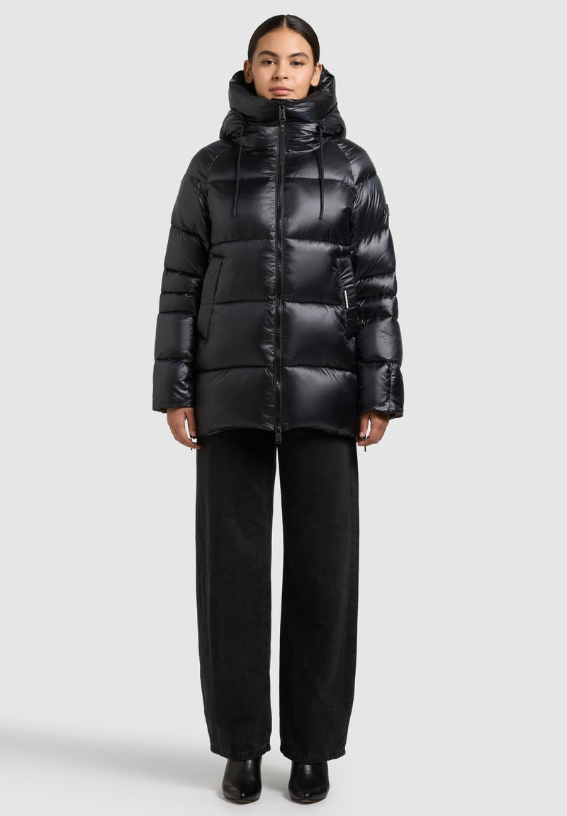 Chaqueta acolchada negra con cuello alto, textura brillante, cremallera frontal y capucha con cordón. Lleva puesta sobre pantalones negros de pierna ancha y botas de tacón.