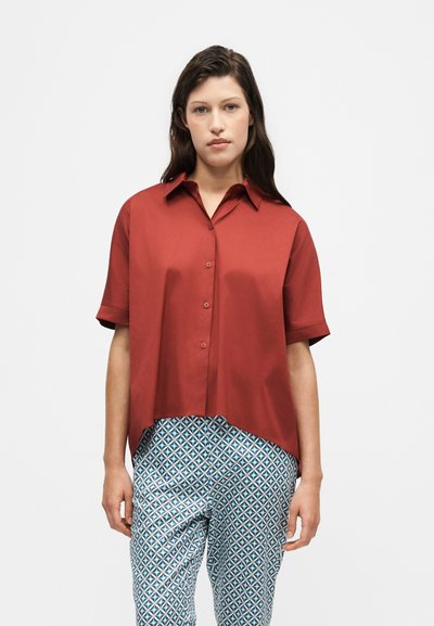 Femme portant une chemise ample à manches courtes couleur rouille boutonnée et un pantalon bleu et blanc à motifs, debout sur un fond uni.