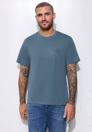 Mann mit kurzen Haaren und Tätowierungen auf beiden Armen, der ein blaues T-Shirt und blaue Jeans trägt, steht vor einem schlichten weißen Hintergrund.