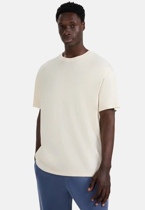 Kremowy t-shirt z krótkim rękawem o okrągłym dekolcie, luźnym kroju i gładkiej fakturze, zestawiony z niebieskimi spodniami.