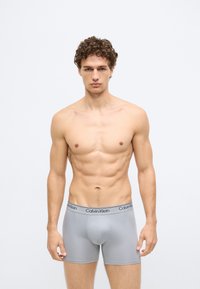 Cuecas boxer cinzentas com cós preto e logo Calvin Klein, desenhadas com um formato ajustado e textura suave, usadas por uma figura masculina.