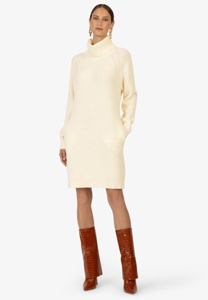 Robe en tricot côtelé crème avec un col roulé haut et des poches latérales, associée à des bottes brillantes en croûte de cuir marron jusqu'au genou.