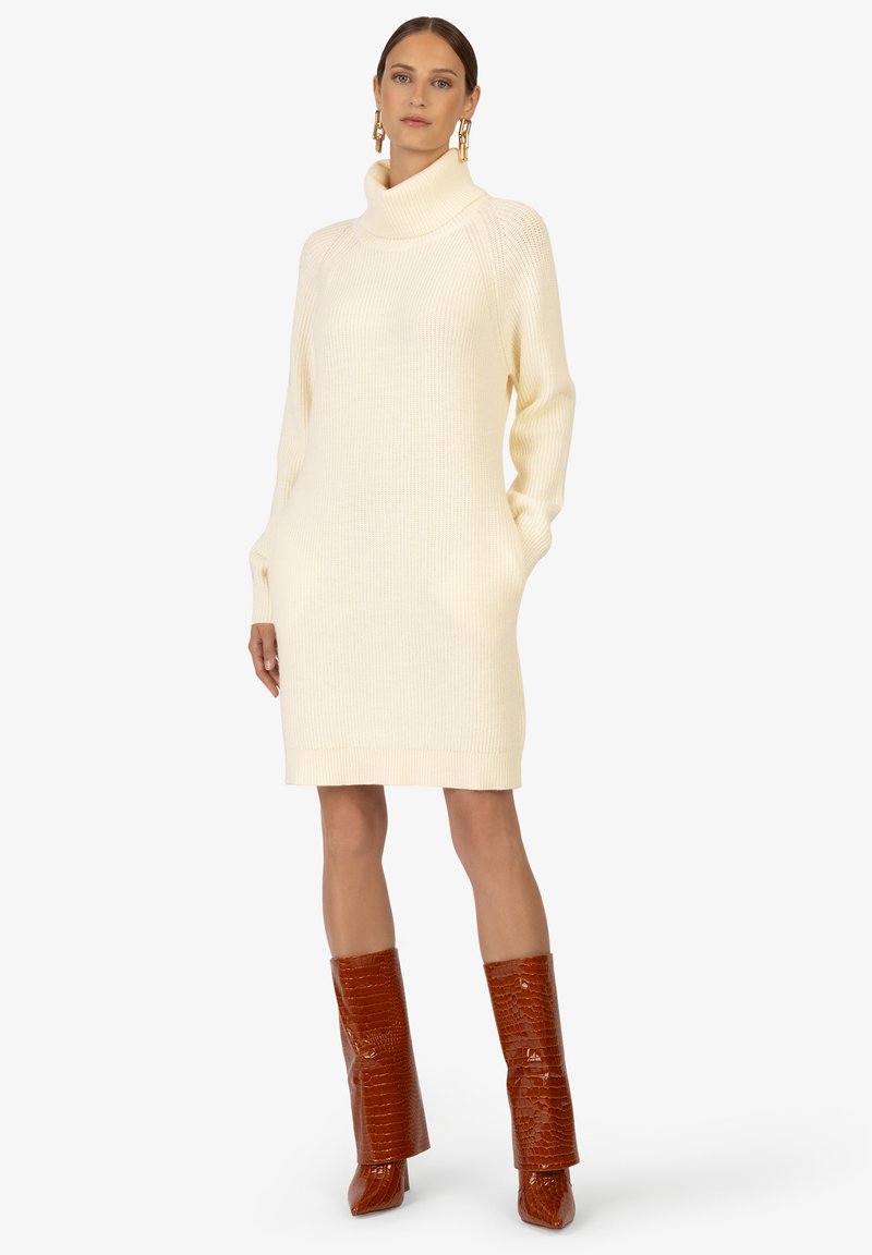 Crème rib gebreid trui-jurk met een hoge col en zijzakken, gecombineerd met glanzende, kniehoge bruine krokodillenleder-embossed laarzen.