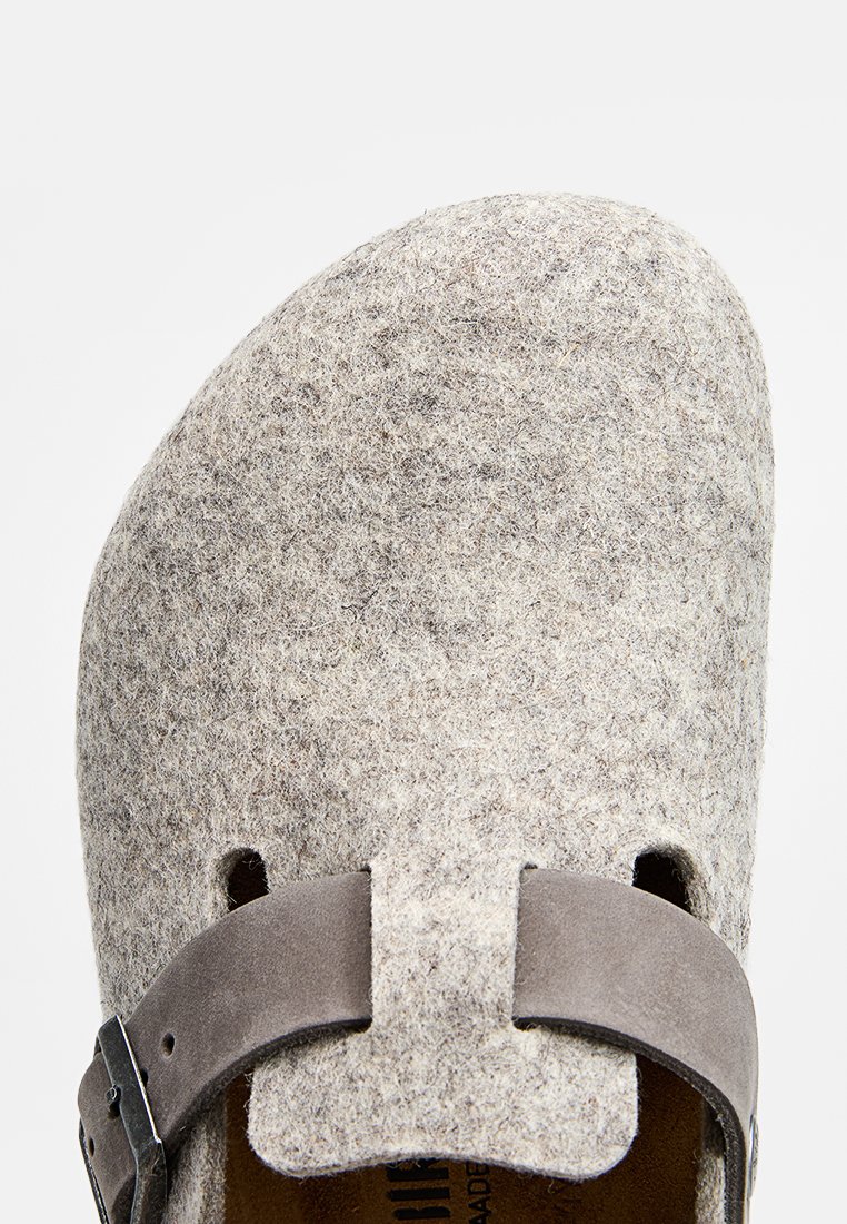 Graue Clogs aus Filz mit abgerundetem Zehenbereich, ausgestattet mit einem verstellbaren braunen Wildlederriemen und seitlichen Ausschnitten für Belüftung. Strukturierte Oberfläche sichtbar.