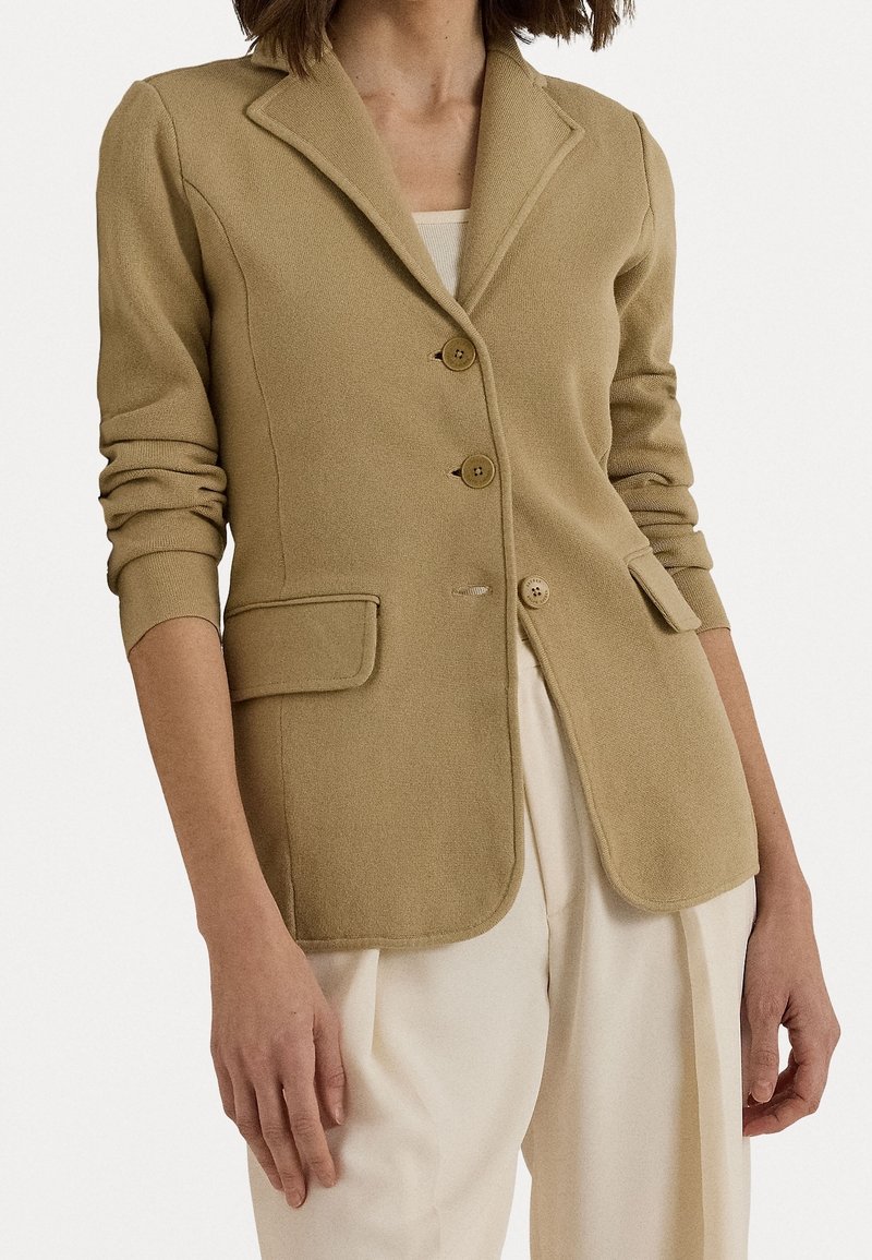 Vaatteet beige teksturoidulla bleiserillä, jossa on notch-läpät, kolme nappia ja kaksi etupussia, yhdistettynä vaaleisiin housuihin.