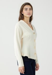 Cremfarbener Cardigan mit V-Ausschnitt, langen Ärmeln und fünf goldfarbenen Knöpfen auf der Vorderseite. Weiche Textur und entspannte Passform. Über dunklen Jeans getragen.
