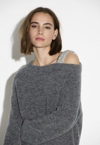 Grijze, oversized trui met een zachte textuur en een off-shoulder ontwerp. Bevat een metalen, glinsterende schouderbandaccent aan één schouder.