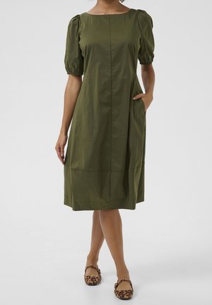 Woman wearing knee-length olive green dress with puff sleeves and leopard print flat shoes, standing with one hand in pocket.