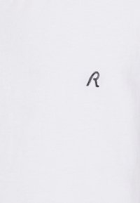 Biely bavlnený tričko s malým čiernym vyšitým písmenom "R" na ľavej hrudi. Tkanina má hladkú textúru.