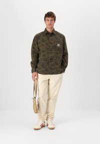 Camicia a maniche lunghe con motivo camouflage in tessuto verde scuro, con tasca a toppa. Abbinata a pantaloni in corduroy di un beige chiaro e sneakers bianche con strisce rosse.