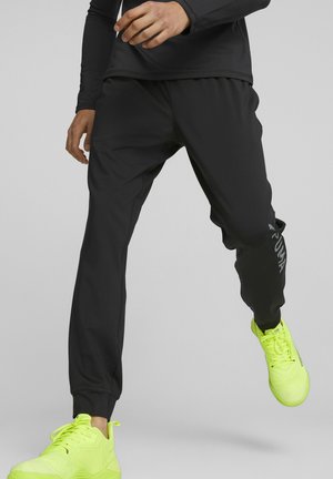 Person iført sorte Puma træningsbukser og klare gule sneakers i en midtgående positur mod en ensfarvet baggrund.