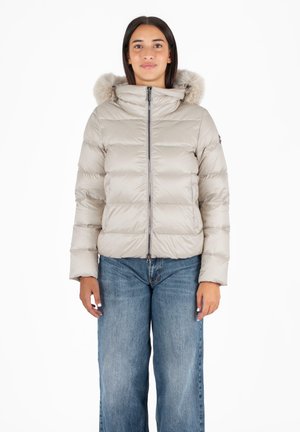 Giacca beige gonfiabile con cappuccio bordato in pelliccia, zip frontale e pannelli cuciti. Abbinata a jeans a gamba larga di colore blu chiaro.