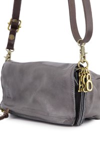 Grijze leren schoudertas met een gestructureerde afwerking. Voorzien van een bruine strap, messing beslag en decoratieve metalen bedels aan de rits.