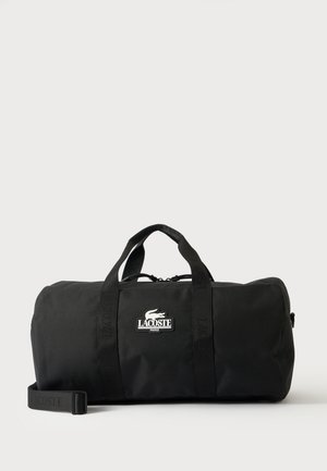 Sac de sport cylindrique noir Lacoste avec deux poignées, une bandoulière amovible, un logo à l’avant et une fermeture éclair sur le dessus.