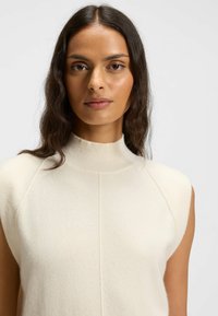 Knit sleeveless turtleneck top in light cream, featuring a ribbed collar, smooth texture, and subtle seam detailing on the shoulders.