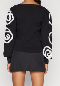 Black sweater with white fluffy spiral patterns on sleeves, ribbed cuffs and hem, relaxed fit, paired with a dark skirt.