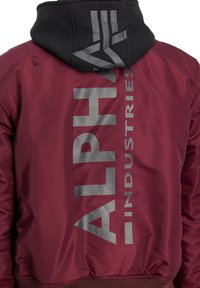 Burgunderfarbene Bomberjacke mit schwarzer Kapuze, mit großen reflektierenden Text auf dem Rücken und gerippten Bündchen. Glattes, glänzendes Material.