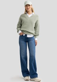 Lichtgroene gebreide trui met een v-hals, gedragen over een witte overhemd met kraag. Gepaard met wijde blauwe jeans en beige sneakers.