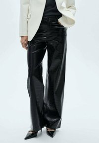 Zwarte wijde broek van glanzend faux leer met een krokodillenpatroon, gecombineerd met zwarte puntige hakken en een witte blazer.