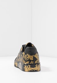 Černá teniska se zlatým zdobeným vzorem, hladkým koženým svrškem, kulatou špičkou a plochou gumovou podrážkou. Na patě se nachází značka Versace Jeans Couture.