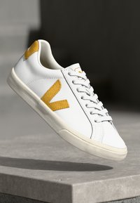 Valged tossud, mis on valmistatud siledast nahast kollase tekstuuriga V-logoga, valgete paelte ja kummitaldadega, omades minimalistlikku disaini ja puhtaid jooni.