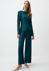 Top lungo a maniche lunghe plissettato color teal e pantaloni a gamba larga con una texture a coste. Abbinati a scarpe con tacco nude.