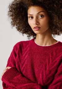 Pūkuotas, tamsiai raudonas megztinis su briaunotu apykaklės kraštu ir zigzago raštu. Ilgos, plačios rankovės ir tekstūruota audinio struktūra suteikia šilumą ir komfortą.