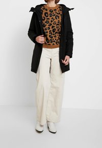 Czarna parka z kapturem nałożona na brązowy sweter w cętki lamparta, w zestawieniu z beżowymi szerokimi spodniami z korduroju i białymi botkami do kostki.