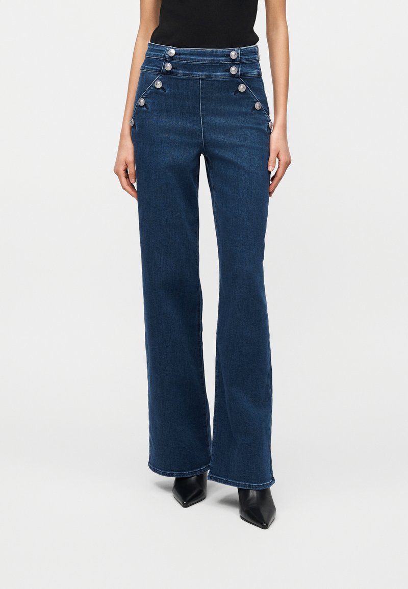 Γυναίκα που φοράει ψηλόμεσα σκουρόχρωμα μπλε τζιν με ασημένια κουμπιά κατά μήκος της μέσης και των γοφών, συνδυασμένα με μαύρα μποτάκια με μυτερή μύτη.