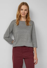 Vrouw met lichtbruin haar draagt een grijs en zwart gestreepte top met driekwartmouwen en donkere rode broeken, staand tegen een effen lichte muur.