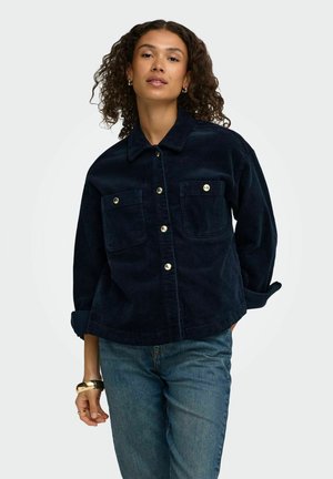 Femme aux cheveux bouclés portant une veste en velours côtelé bleu foncé avec des boutons dorés et un jean bleu, debout devant un fond clair uni.